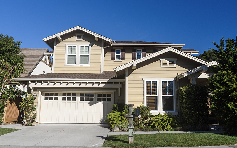 Two story home, painted a tan color with blue sky behind it and trees on either side.
