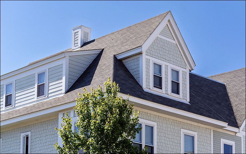 Two story home, painted a tan color with blue sky behind it and a tree in the foreground.