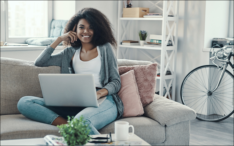 Woman smiling on couch in living room using laptop. Bike and bookshelf behind her.