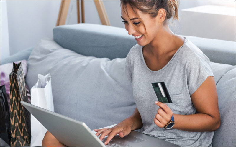 Woman smiling while sitting on couch using credit card to make a purchase on laptop.