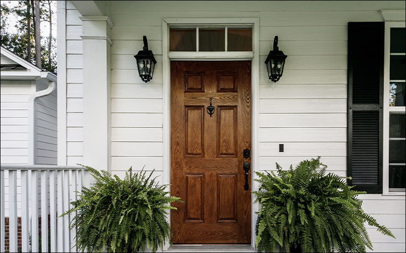 Wooden front door on white house with two plants on either side.