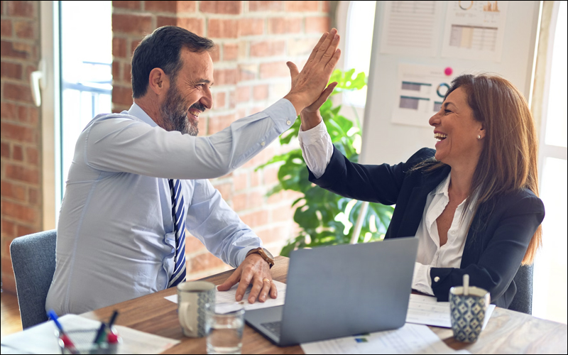 Man and woman doing a high five and smiling at a table with a laptop and paperwork in front of them.